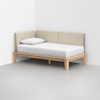 A minimalist daybed with a light wood frame, beige upholstered backrest, white mattress, and a single white pillow, placed in the corner of a bright, empty room with light gray flooring.