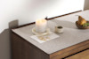 A lit white candle on a saucer sits atop a dresser with a postcard, a small bowl with a matchstick, and a bowl containing a pear and a kiwi, all in soft natural light.