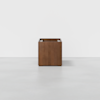 A square, wooden planter box with a smooth, brown finish sits centered on a light gray floor against a plain white background. The planter is empty and has a modern, minimalist design.