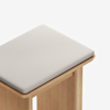 A close-up of a minimalist wooden stool with a light-colored cushioned seat and smooth edges, set against a plain white background.