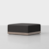 A modern, square ottoman with a dark upholstered cushion and a light wood base, set against a plain white background.