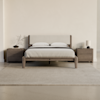 Minimalist bedroom with a wooden bed frame, beige upholstered headboard, two pillows, and matching nightstands on each side. One nightstand has a vase with branches, the other holds a lamp and books. A light rug covers the floor.