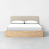 A modern bed with a light beige upholstered headboard, white bedding, two pillows, and a light wood frame, set against a plain white wall and a gray floor.