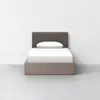 A minimalist twin bed with a brown upholstered headboard, white bedding, and a light wood frame, set against a plain white wall and gray floor.