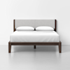 A modern bed with a dark wooden frame and a light gray upholstered headboard, neatly made with white bedding and two white pillows, set against a plain white wall.