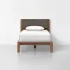 A single wooden bed with a dark upholstered headboard, white bedding, and a pillow, placed against a plain white wall on a light gray floor.