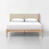 A modern wooden bed frame with a beige upholstered headboard, white bedding, and two white pillows, set against a plain white wall on a light gray floor.
