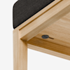 Close-up of a wooden bench corner with a light wood finish and a black cushion on top; a round black button or switch is visible underneath the seat.