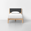 A minimalist single bed with a light wood frame, dark gray upholstered headboard, and white bedding with a pillow, placed against a plain white wall on a light gray floor.
