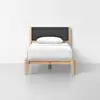 A minimalist single bed with a light wood frame, dark gray upholstered headboard, and white bedding with a pillow, placed against a plain white wall on a light gray floor.