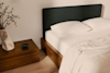A neatly made bed with white bedding and two pillows next to a wooden nightstand holding a sculptural vase with green branches and a small dark bowl, set against a light-colored wall and wood floor.
