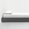 A minimalist bed with a gray upholstered frame, white bedding, and a single white pillow, placed against a plain white wall on a light-colored floor.