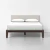 A modern bed with a dark wooden frame, beige upholstered headboard, and white bedding with two pillows, set against a plain white wall and floor.