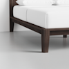 Close-up view of a dark wooden bed frame with clean lines, white bedding, and white pillows, placed on a light gray floor near a white wall.