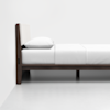 A minimal wooden bed frame with a padded headboard, neatly made with white sheets and a pillow, positioned against a white wall on a white floor.