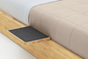 A close-up of a wooden bed frame with a small, rectangular, black felt tray attached to its side, next to a beige quilted comforter and white sheet on the bed.