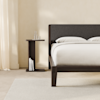 A modern bedroom with a dark wood bed frame and gray upholstered headboard, white bedding, a matching side table holding a carafe and glass, books on the lower shelf, light curtains, and a neutral rug.