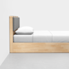Side view of a minimalist wooden bed with a padded gray headboard, white pillow, and neatly made white bedding, placed against a plain white wall on a smooth gray floor.
