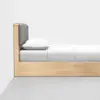 Side view of a minimalist wooden bed with a padded gray headboard, white pillow, and neatly made white bedding, placed against a plain white wall on a smooth gray floor.