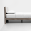 Side view of a minimalist wooden bed frame with a light-colored padded headboard, white pillow, and white bedding in a bright, simple room with white walls and floor.