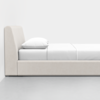 A minimalist beige upholstered bed with a white mattress, white bedding, and a white pillow, positioned against a plain white wall on a light gray floor.