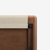 Close-up of a wooden bed frame corner with a light beige fabric panel attached at the top, showing smooth rounded edges and dark hardware details.