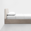 A minimalist bed with a light wood frame, a beige upholstered headboard, a white mattress, white bedding, and a white pillow, positioned against a plain white wall on a gray floor.