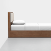 A minimalist wooden bed with a rectangular headboard featuring beige cushions, neatly made with white bedding and pillows, set against a plain white wall and gray floor.