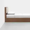 A minimalist wooden bed with a rectangular headboard featuring beige cushions, neatly made with white bedding and pillows, set against a plain white wall and gray floor.