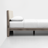 A minimalist wooden bed frame with a light beige upholstered headboard, white bedding, and a single white pillow, set against a plain white wall and floor.