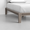 Close-up of a modern wooden bed frame with light brown legs, white bedding, and a pillow, set against a light gray floor and white wall.