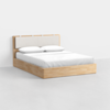 A modern wooden bed frame with a light wood finish, featuring a padded beige headboard. The bed is neatly made with white sheets, a white blanket, and two white pillows. The background is plain and minimalistic.