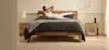 A woman stretches and smiles while sitting up in a neatly made bed with dark gray bedding in a bright, minimalist bedroom with wooden furniture and soft natural light.