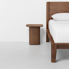 A modern wooden bed frame with white bedding is positioned next to a matching wooden nightstand, set against a plain white wall and light gray floor.