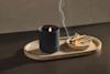 A black candle in a matte holder burns on an oval stone tray next to a small round dish with several burnt matchsticks, with smoke rising from the matches. The background is neutral and minimal.