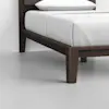 A close-up view of a wooden bed frame with crisp white bedding and a pillow, positioned on a smooth, light grey floor against a plain white wall.