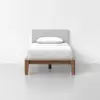 A single bed with a wooden frame, white bedding, and a light gray upholstered headboard, positioned against a plain white wall in a minimalistic room.