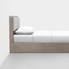 A minimalist wooden bed with a padded headboard, white bedding, and a single white pillow, positioned against a plain white wall on a light gray floor.