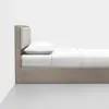 A minimalist wooden bed with a padded headboard, white bedding, and a single white pillow, positioned against a plain white wall on a light gray floor.