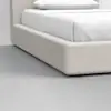 Close-up of a modern beige upholstered bed frame with white bedding and pillow, set on a smooth, light gray floor in a minimalist room.