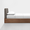 A minimalist bed with a wooden base, a gray upholstered headboard, and white bedding, placed against a plain white wall on a light gray floor.