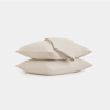 Two beige pillows stacked on top of each other with a matching folded bedsheet draped over the top pillow, set against a plain white background.