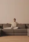 A person sits relaxed on a large gray sectional sofa, dressed in light-colored, casual clothing, against a plain beige wall and hardwood floor.