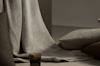 A neutral-toned scene with a textured beige blanket draped over a chair, soft gray and brown cushions on the floor, and a small, translucent brown glass placed beside them.