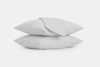Two white pillows are stacked on top of each other against a plain white background. A folded white sheet or pillowcase is draped neatly over the top pillow.