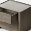 A modern, light wood nightstand with clean lines is shown against a plain white background. One drawer is partially open, revealing its interior. The furniture has a minimalist and boxy design.