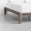A close-up view of a wooden bed frame with clean lines and a modern design, featuring a white mattress and pillows on top, set against a light gray floor and white wall.