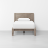 A minimalist wooden twin bed with a rectangular headboard, white bedsheet, and a single white pillow, set against a plain white wall and light gray floor.
