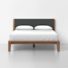 A modern bed with a wooden frame, dark upholstered headboard, white bed linens, and two white pillows, set against a plain white wall and floor.