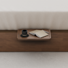 A wooden tray on a wooden bed frame holds a cup on a saucer, an open notebook, and a pen, next to a neatly made bed with white bedding.